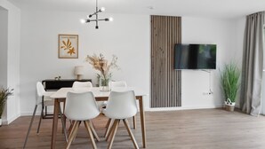 A modern living room with a dining area, featuring a flat-screen TV, a wooden floor, and a white wall with a framed artwork. - Modern KüstenHygge 2 with garden & parking space (Westerdeichstrich)