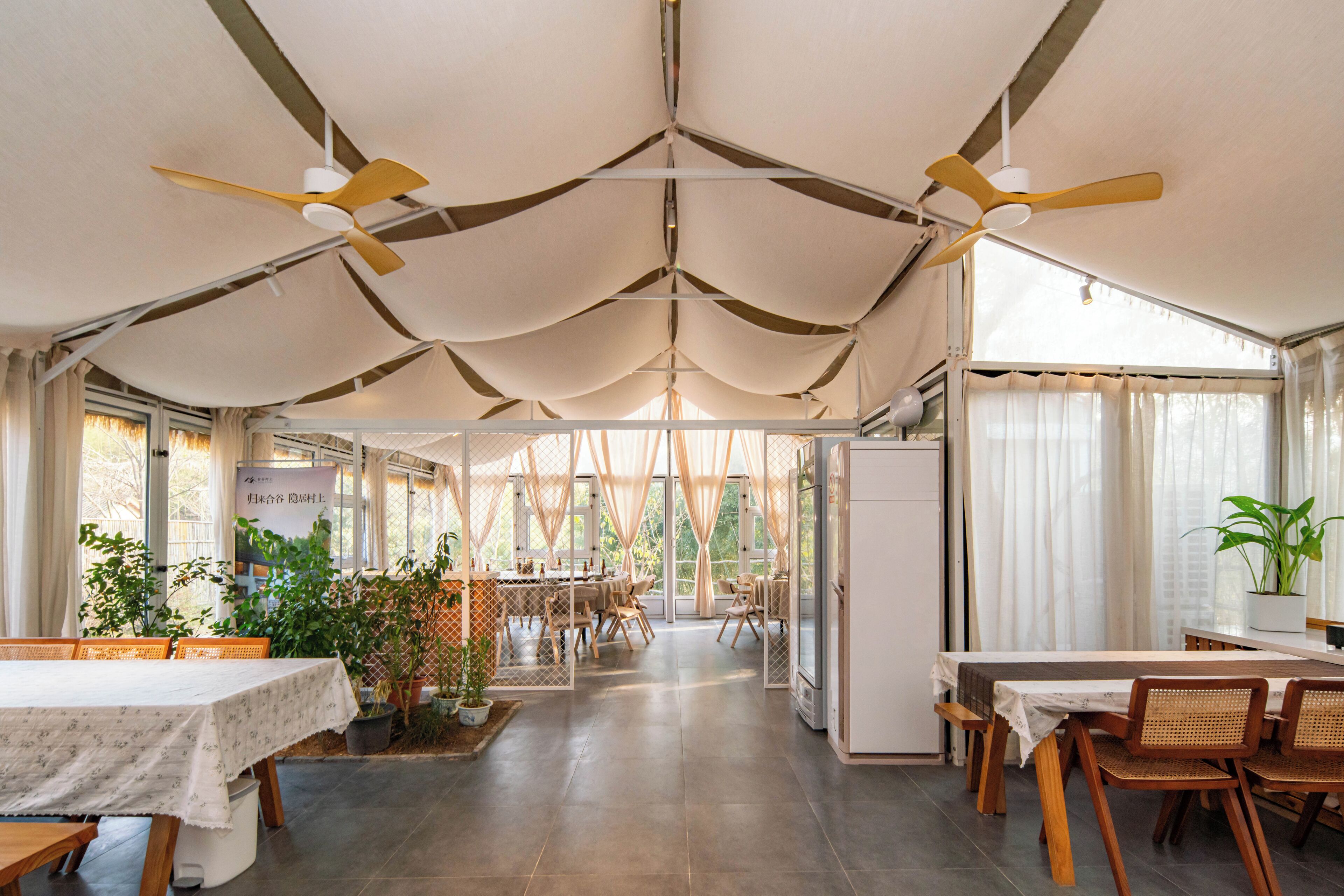 Une salle à manger spacieuse avec de hauts plafonds, des ventilateurs de plafond et de grandes fenêtres. Des tables sont disposées avec des chaises, et des plantes en pot sont placées un peu partout dans la pièce.