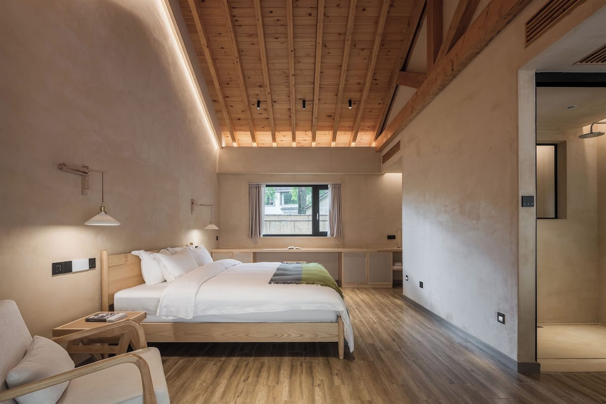A bedroom with a wooden ceiling, a bed with white bedding, a window, and a desk.