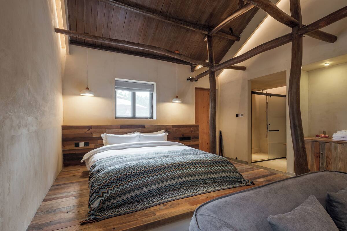 A bedroom with a wooden ceiling, a bed with a patterned bedspread, a grey sofa, and a wooden floor.