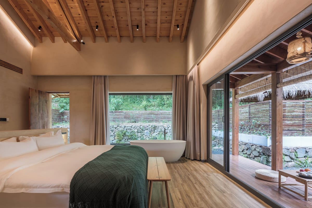 A bedroom with a large bed, a wooden bench, a bathtub, and a view of a stone wall and greenery outside.