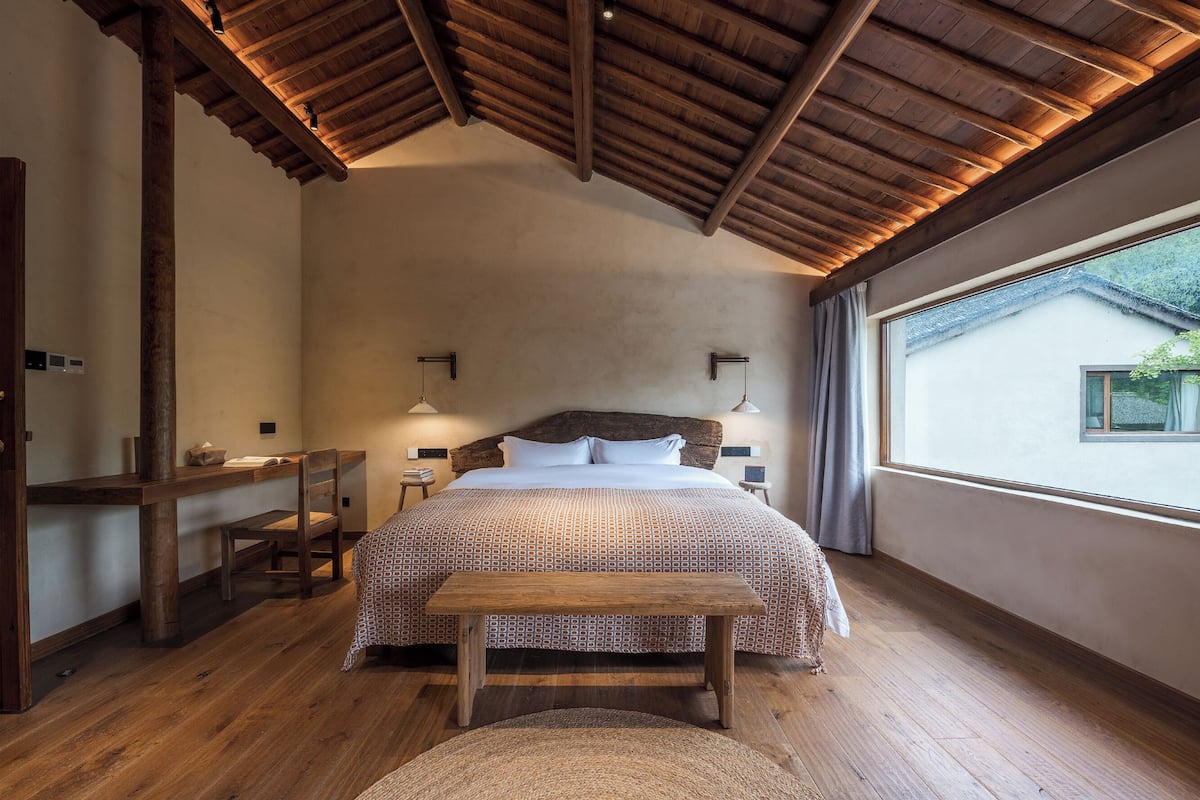 A bedroom with a wooden ceiling, a bed with patterned bedding, a nightstand, a bench, and a view of a house through a window.