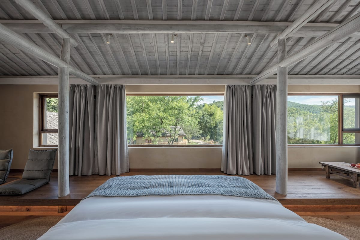 A bedroom with a large bed, wooden beams on the ceiling, and a view of trees through the window.
