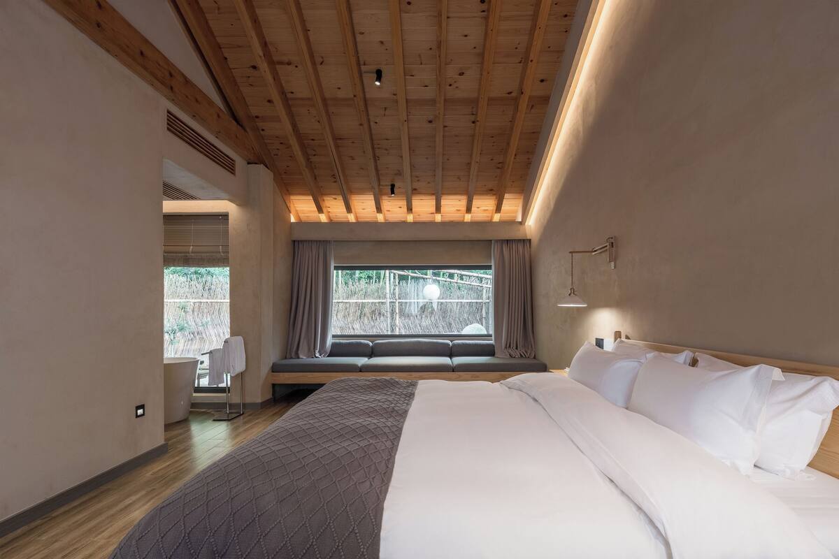 A bedroom with a large bed, a window seat, and a view of greenery outside.
