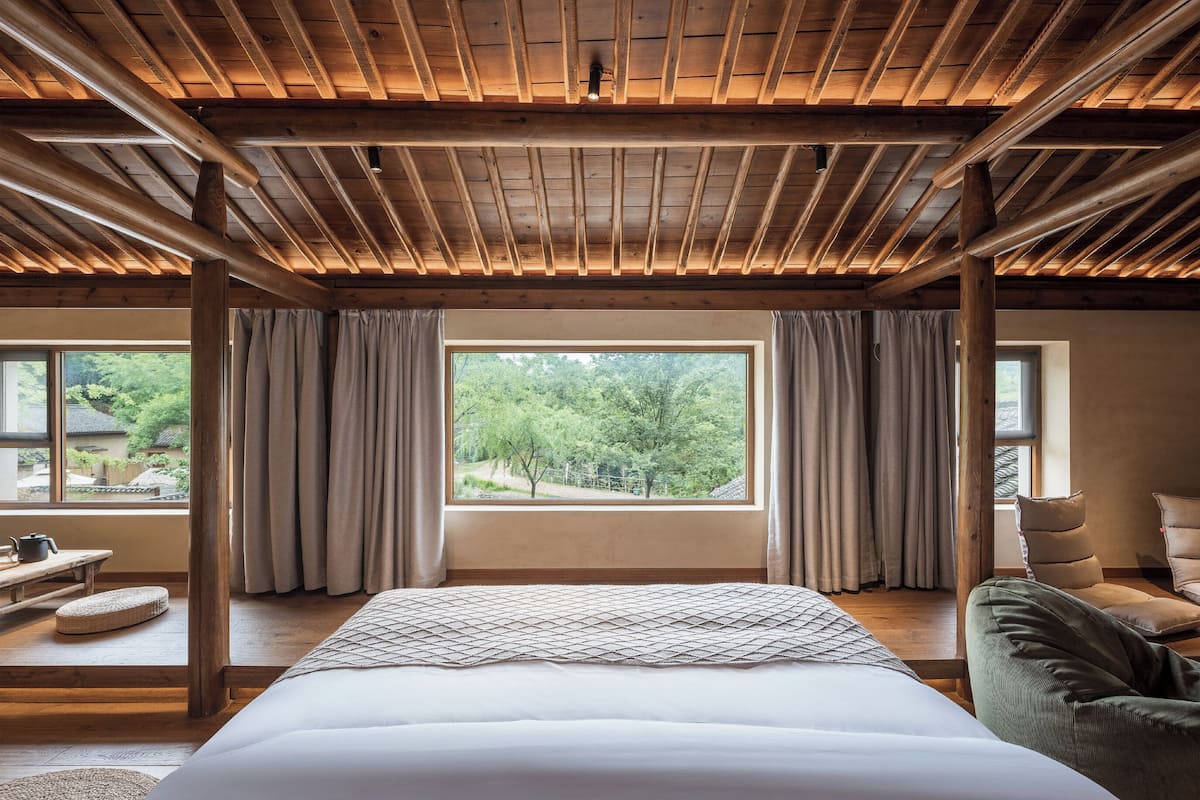 A bedroom with a large bed, wooden ceiling beams, a view of trees, and a window with curtains.
