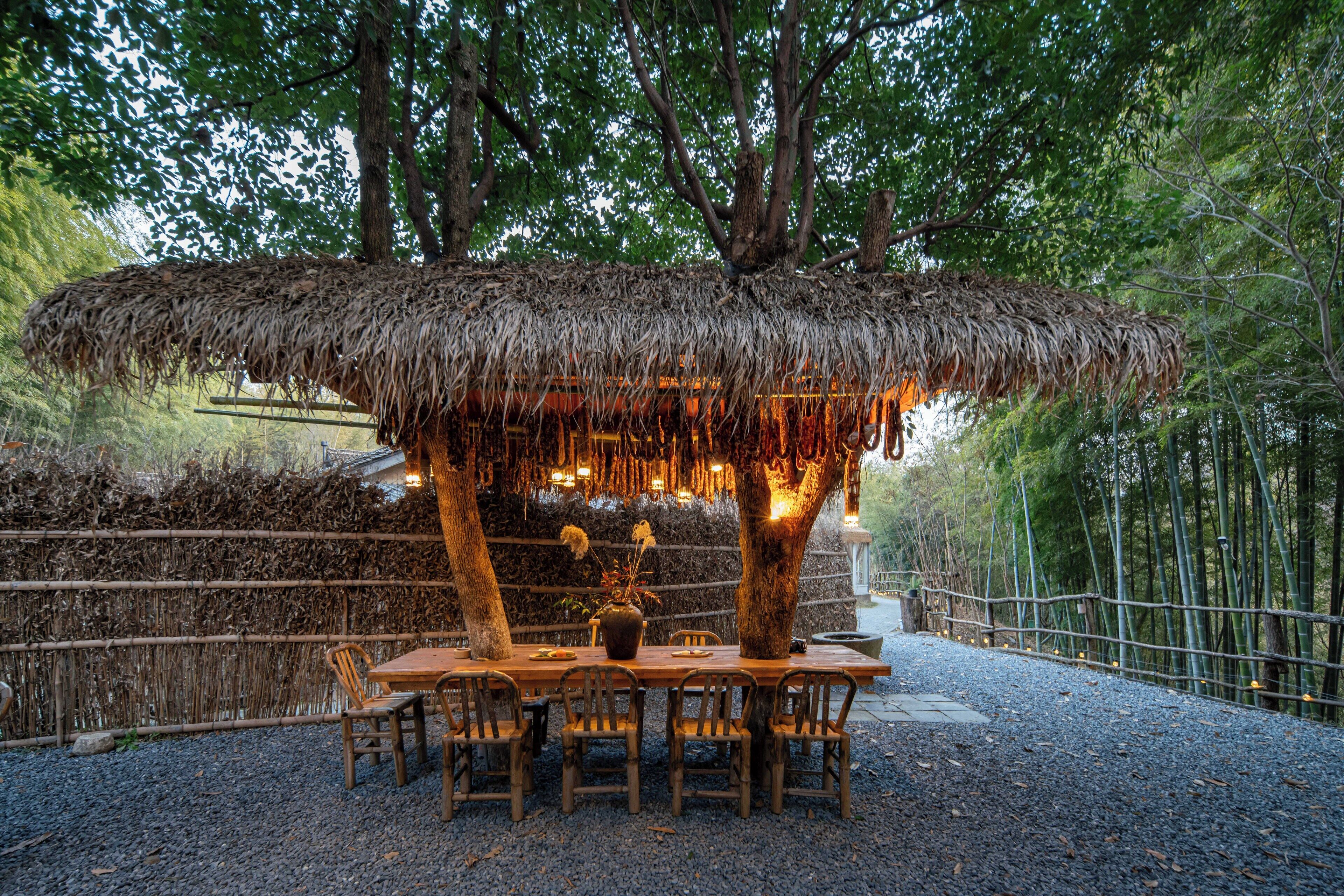 Un espacio de comedor al aire libre con techo de paja, rodeado de árboles y una valla de bambú.