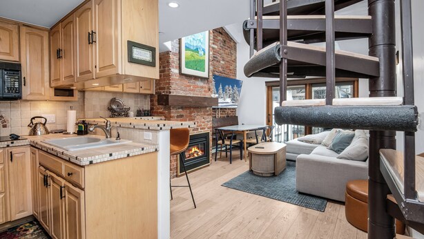 A modern kitchen with wooden cabinets, a central island, and a fireplace. There is a dining area with a table and chairs, and a living room with a sofa and a fireplace.