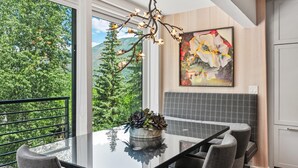 Une cuisine moderne avec une table à manger en verre, des chaises grises, une plante en pot, un luminaire décoratif en forme de branche et un grand tableau abstrait accroché au mur.