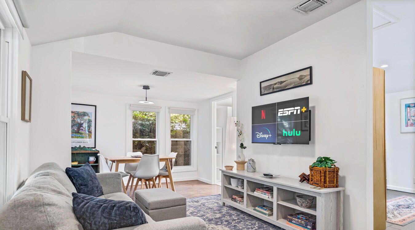 A modern living room with a grey sofa, a dining table with chairs, a TV displaying streaming services, and a bookshelf.