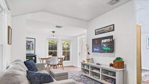 Una sala de estar moderna con un sofá gris, una mesa de comedor con sillas, un televisor mostrando servicios de streaming y una estantería.