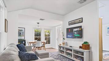 A modern living room with a grey sofa, a dining table with chairs, a TV displaying streaming services, and a bookshelf.