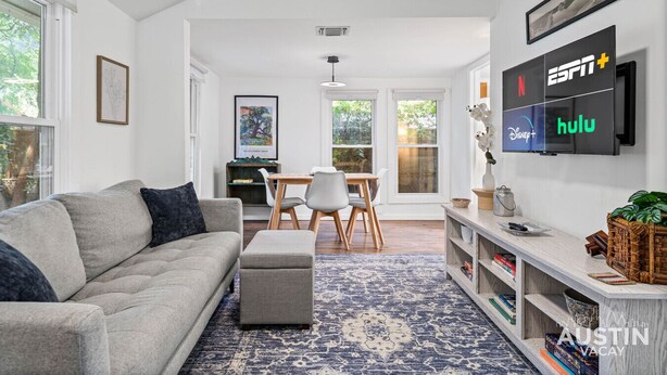 Una sala de estar moderna con un sofá gris, una mesa de centro, un comedor con una mesa y sillas de madera, y un televisor montado en la pared que muestra ESPN+, Hulu y Netflix.