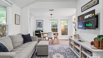 A modern living room with a grey sofa, a coffee table, a dining area with a wooden table and chairs, and a TV mounted on the wall displaying ESPN+, Hulu, and Netflix.