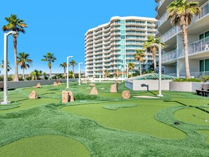 A modern hotel with a curved facade, multiple balconies, and a well-maintained artificial lawn with rock landscaping.