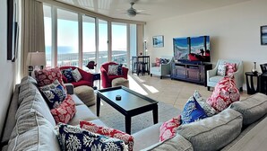 A spacious living room with a large sofa set, a coffee table, and a TV. The room has large windows offering a view of the cityscape. - 3BD/3BA Gulf-Front Condo at Caribe w. Pools, Tennis, Gym, Boat Slips + More (Orange Beach)