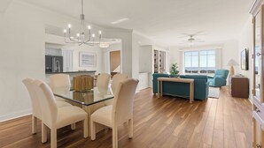 Amplia sala de estar con una mesa de comedor de vidrio, sillas color beige, un sofá verde azulado y una cocina al fondo.