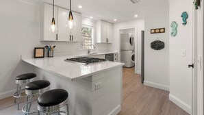 Une cuisine moderne avec des meubles blancs, un îlot central doté de tabourets, un éclairage suspendu et une vue sur la salle à manger.