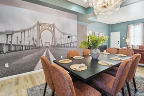 Un coin repas avec une grande table, des chaises en osier, un vase rempli de fleurs et une fresque représentant un pont sur le mur.