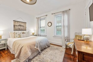Une chambre à coucher avec un grand lit, deux tables de chevet avec des lampes, une commode en bois, un miroir rond et un tableau accroché au mur.