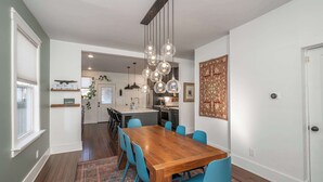 Un comedor con una mesa de madera y sillas azules, una cocina al fondo y una lámpara colgante.