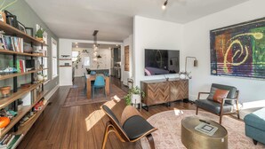 Una sala de estar moderna con suelo de madera, un sofá, una mesa de centro y un televisor. Se observa una estantería y un comedor al fondo.