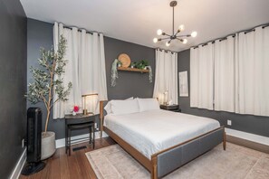 A modern bedroom with a large bed, bedside tables, a floor lamp, a wall-mounted shelf with plants, and a ceiling light fixture. - Northside Nook – Home for 6! (Cincinnati)