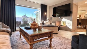 Un salon spacieux doté d’une grande fenêtre, d’un téléviseur à écran plat fixé au mur, d’une table basse en bois et d’un coin salon confortable.
