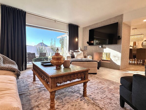 A spacious living room with a large window, a flat-screen TV mounted on the wall, a wooden coffee table, and a comfortable seating area. - Perfect for Every Adventurer! Visit for venues and views (Phoenix)