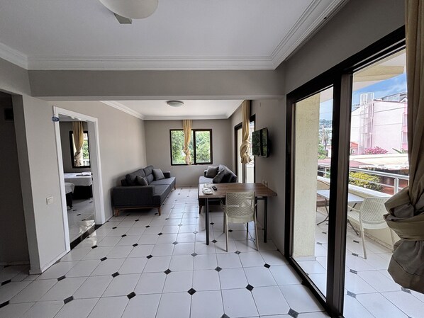 A modern living room with a sofa, a dining table, and a TV. There is a balcony with a view of the city.
