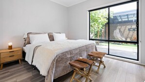 Une chambre avec une grande fenêtre, un lit, une table de chevet et deux tabourets en bois.