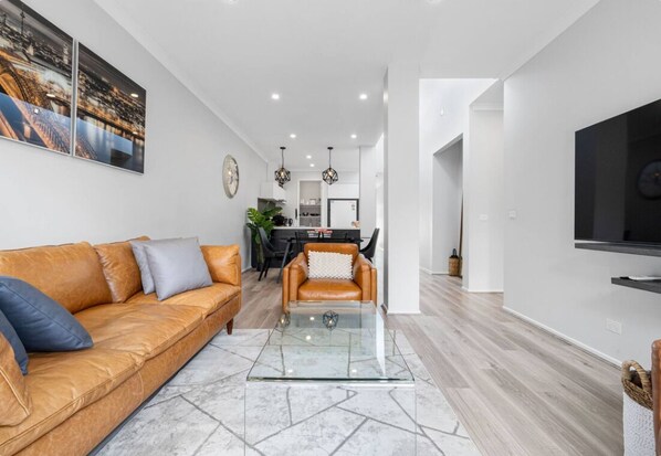 A modern living room with a leather sofa, a glass coffee table, and a dining area in the background.