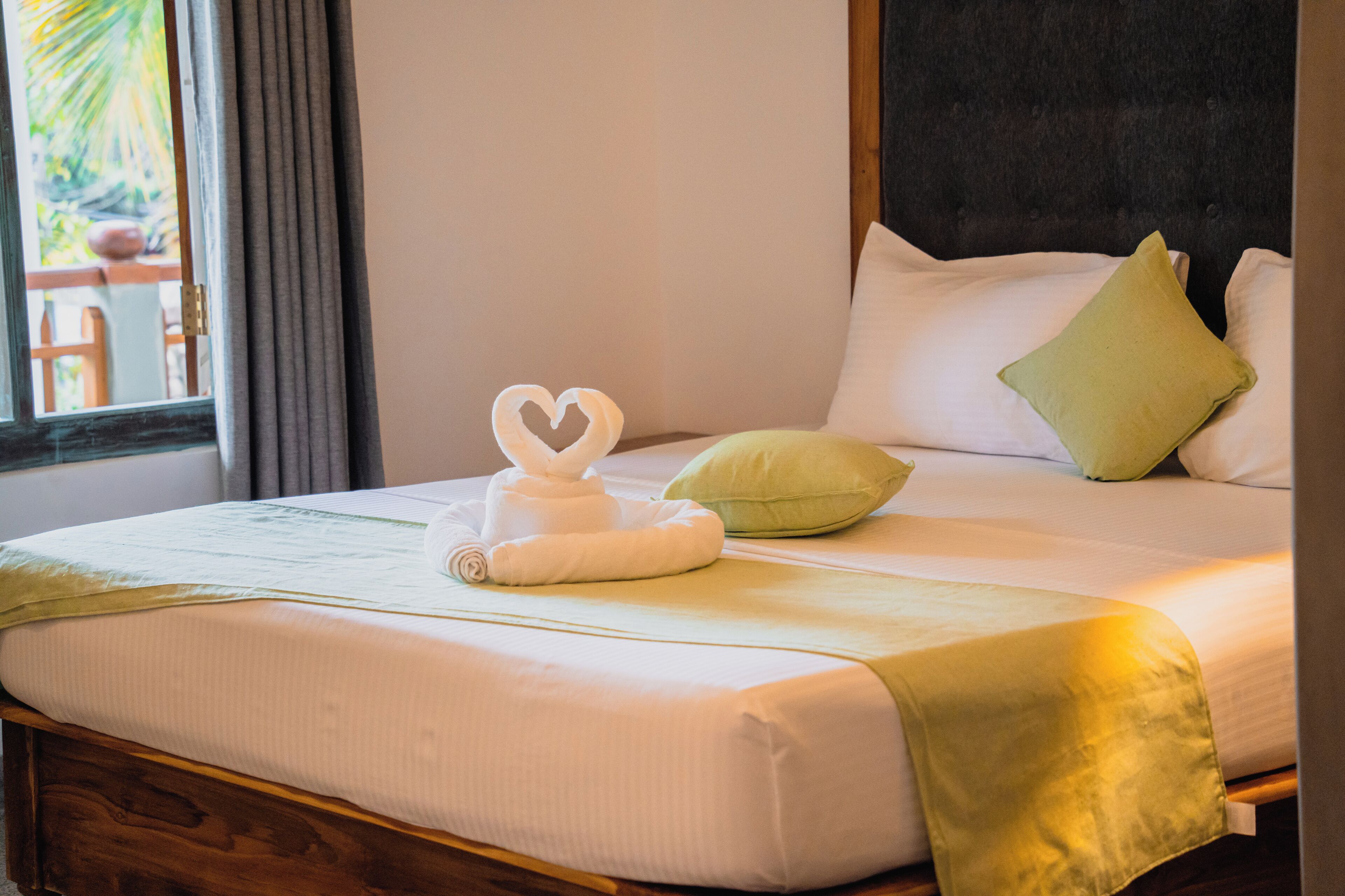 A neatly made bed with a heart-shaped towel, a green pillow, and a wooden headboard.