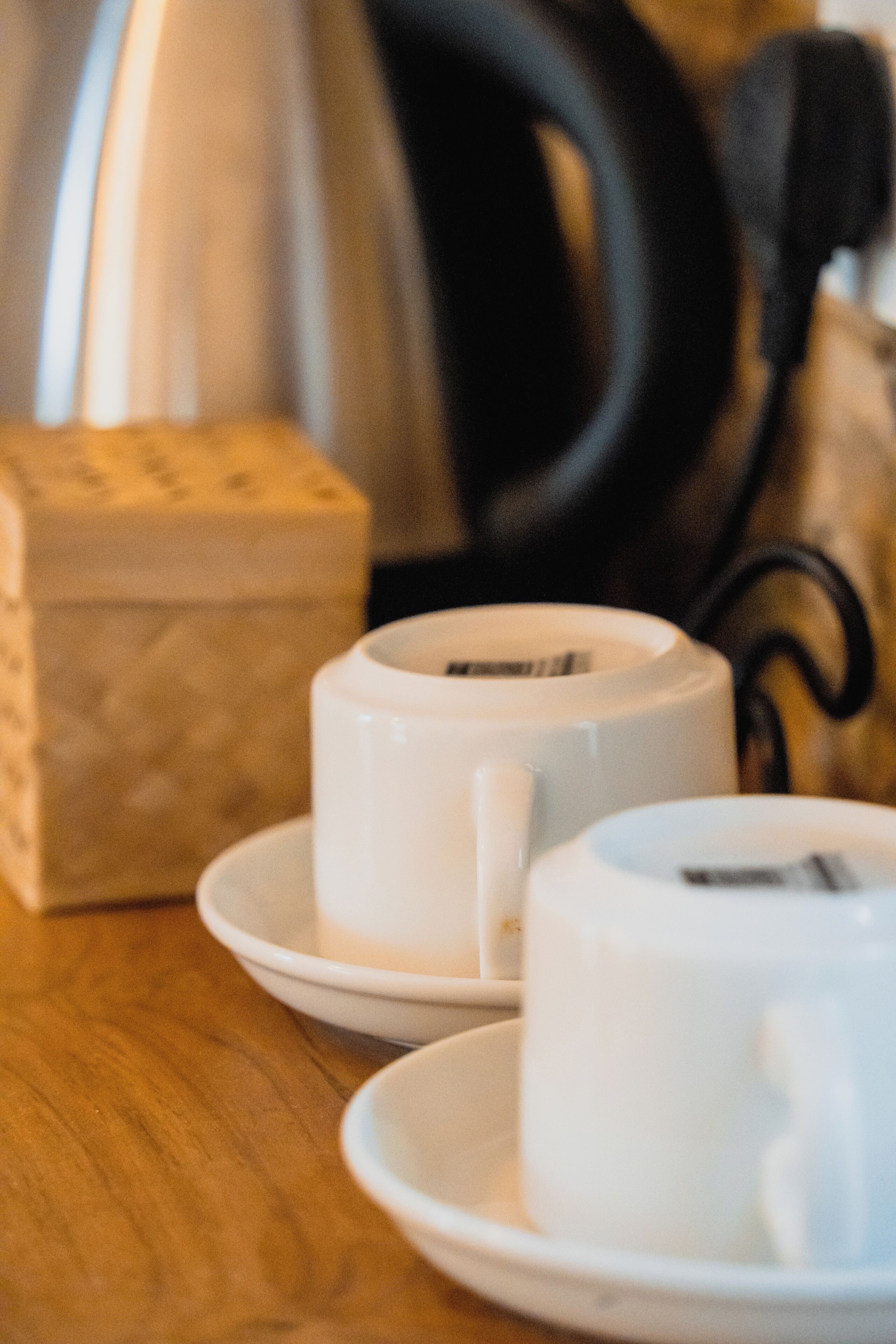 A close-up of a white cup on a saucer, with a blurred background including a black object and a beige box.