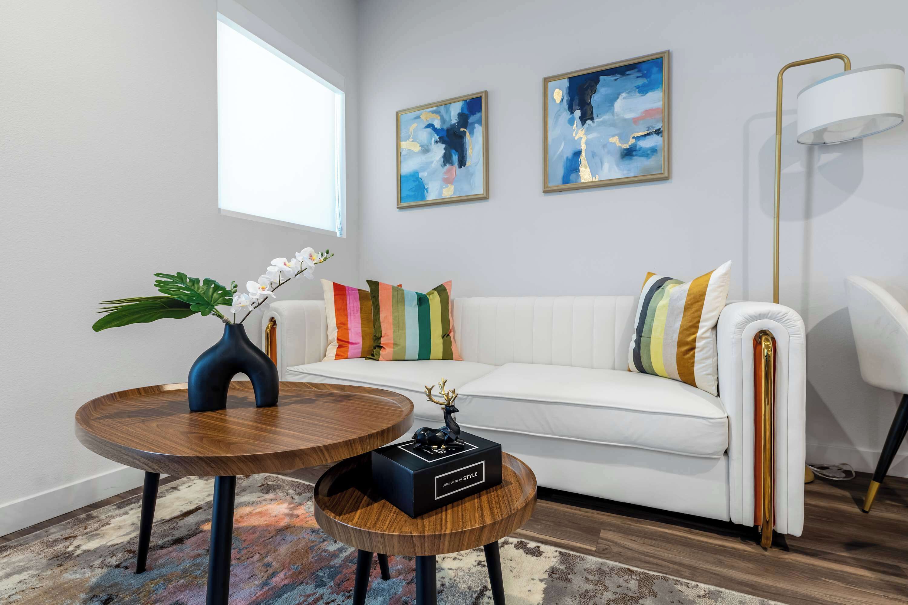 Ein modernes Wohnzimmer mit einem weißen Sofa, bunten Kissen, einem Holztisch, einer Vase mit weißen Blumen und abstrakten Bildern an der Wand.