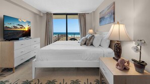 A bedroom with a bed, a TV, a nightstand, a lamp, and a view of the beach. - 🌞Emerald Surf 106: 1st Floor Gulf-Front Condo w/ Pool🌞 (Navarre Beach)