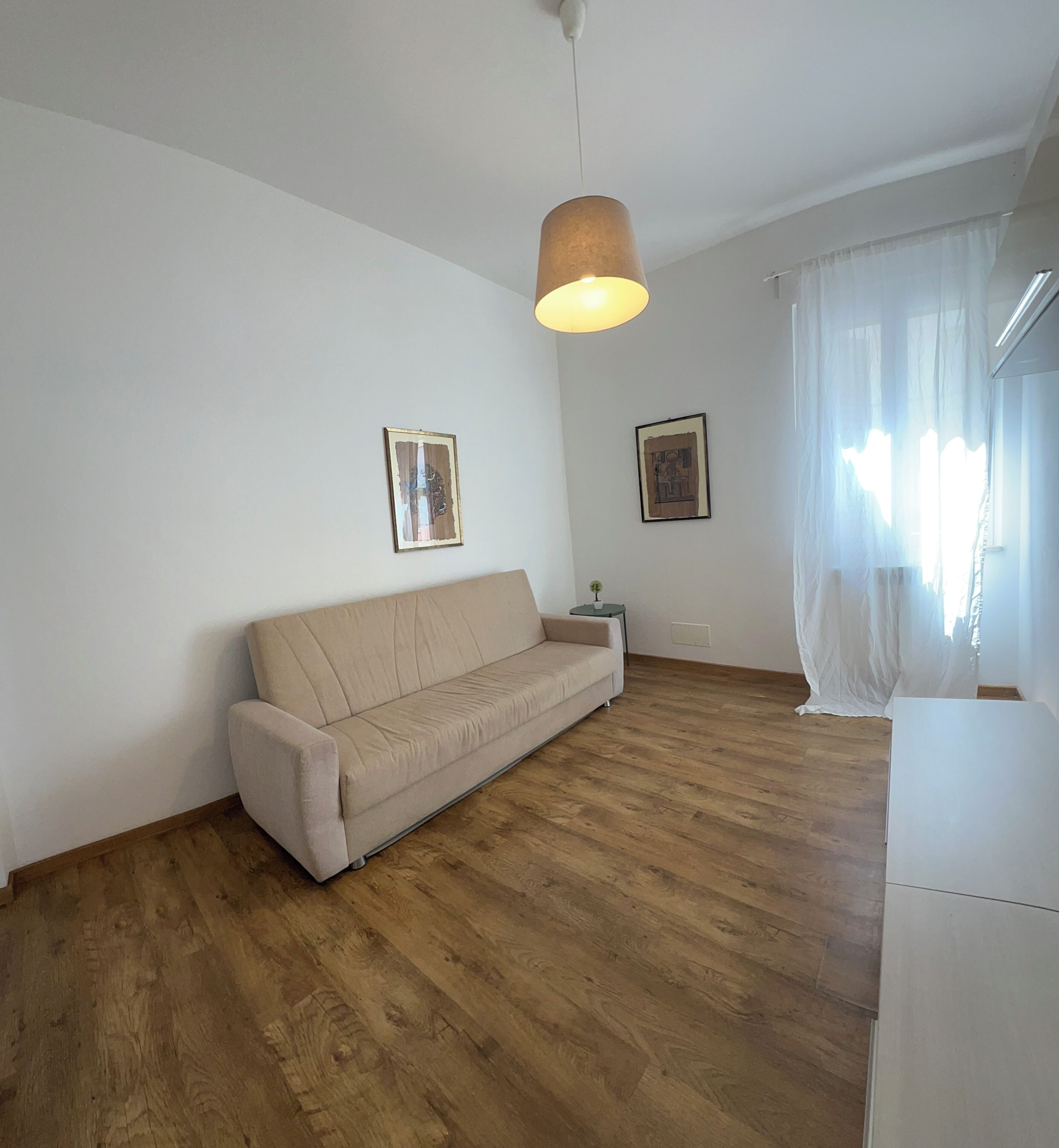 En stue med en beige sofa, trægulv, to billeder i rammer på væggen, en hængende lampe og et vindue med lette gardiner.