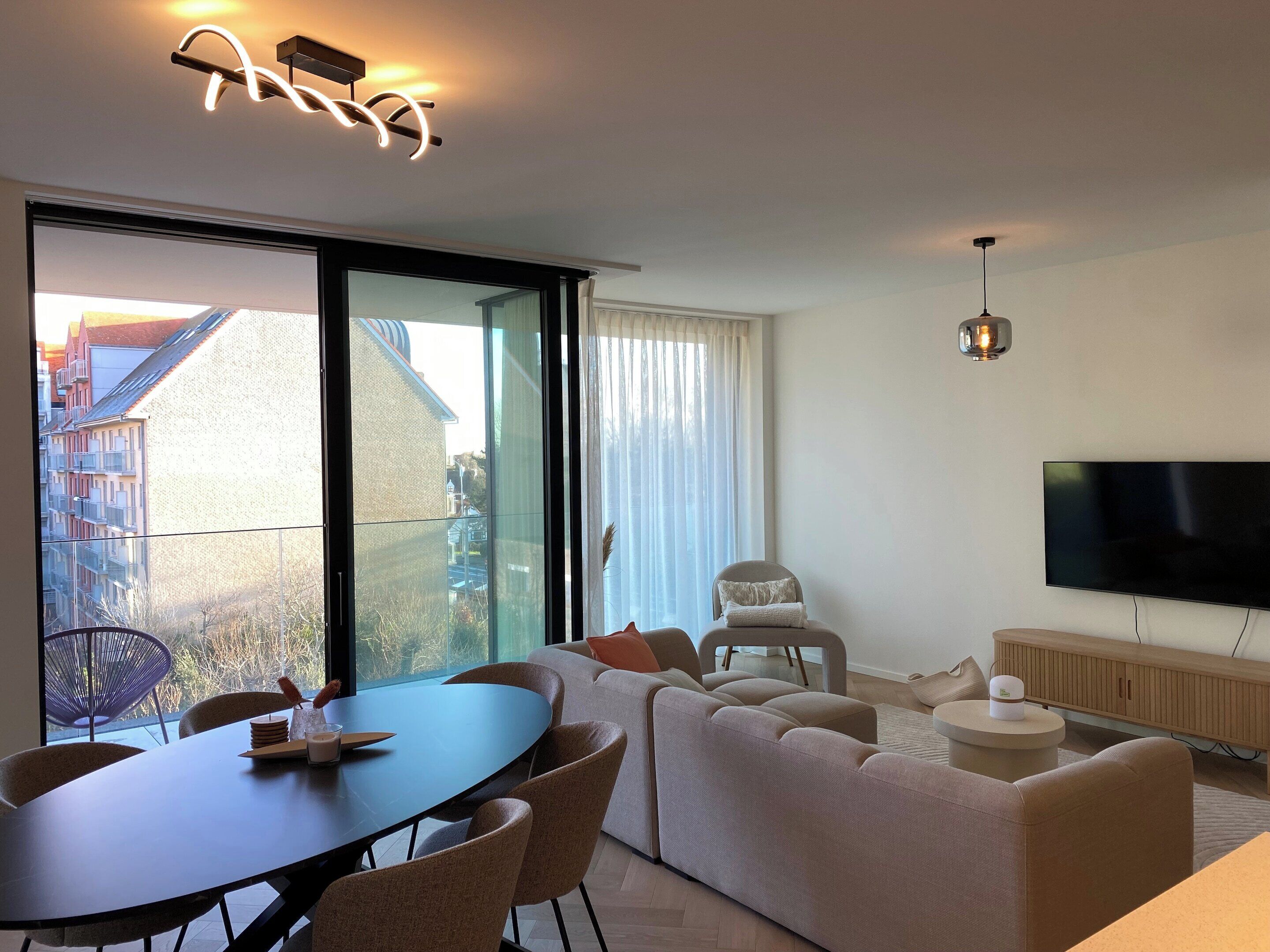 A modern living room with a dining area, large windows, a flat-screen TV, and a cozy sofa.