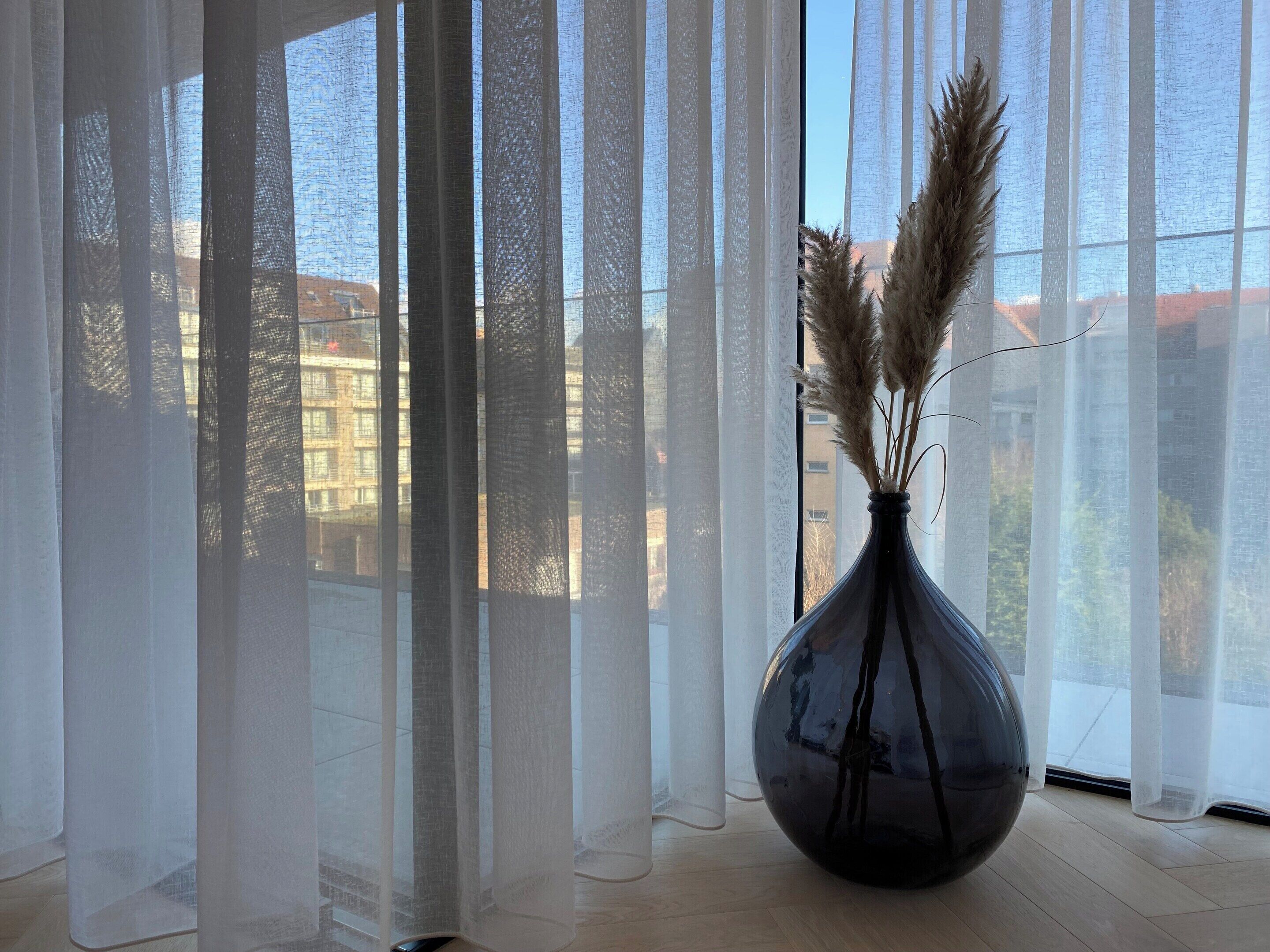 A room with sheer curtains, a vase with pampas grass, and a view of buildings outside.