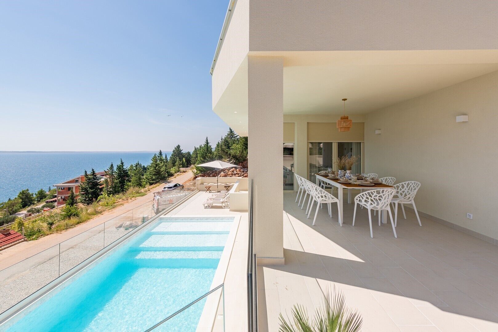 Uma área moderna com piscina ao ar livre e vista clara para o mar, espaço de refeições com cadeiras brancas e uma área de estar coberta.