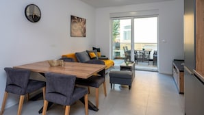 A modern living room with a wooden dining table, grey upholstered chairs, a sofa with yellow cushions, and a large window with a view of the outside.