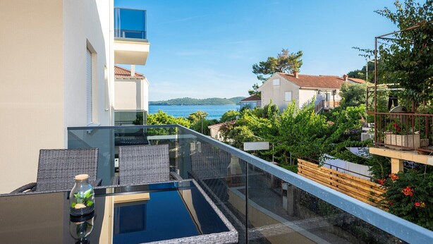 A balcony with a glass barrier, a small table with a plant, and a view of the sea and houses in the background.