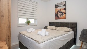 A neatly made bed with white linens and a floral pattern, a bedside table with a lamp, a window with blinds, and a painting of a compass on the wall.