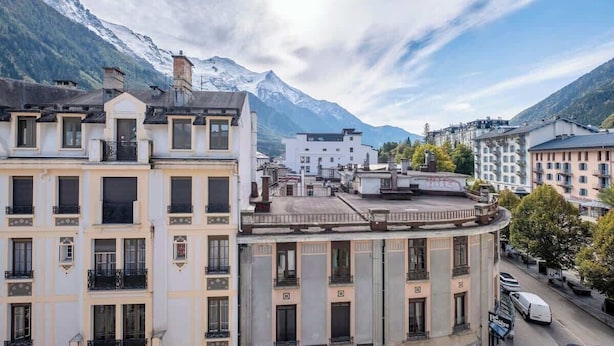 Un edificio con fachada curva, múltiples ventanas y balcones, con montañas y cielo despejado al fondo.