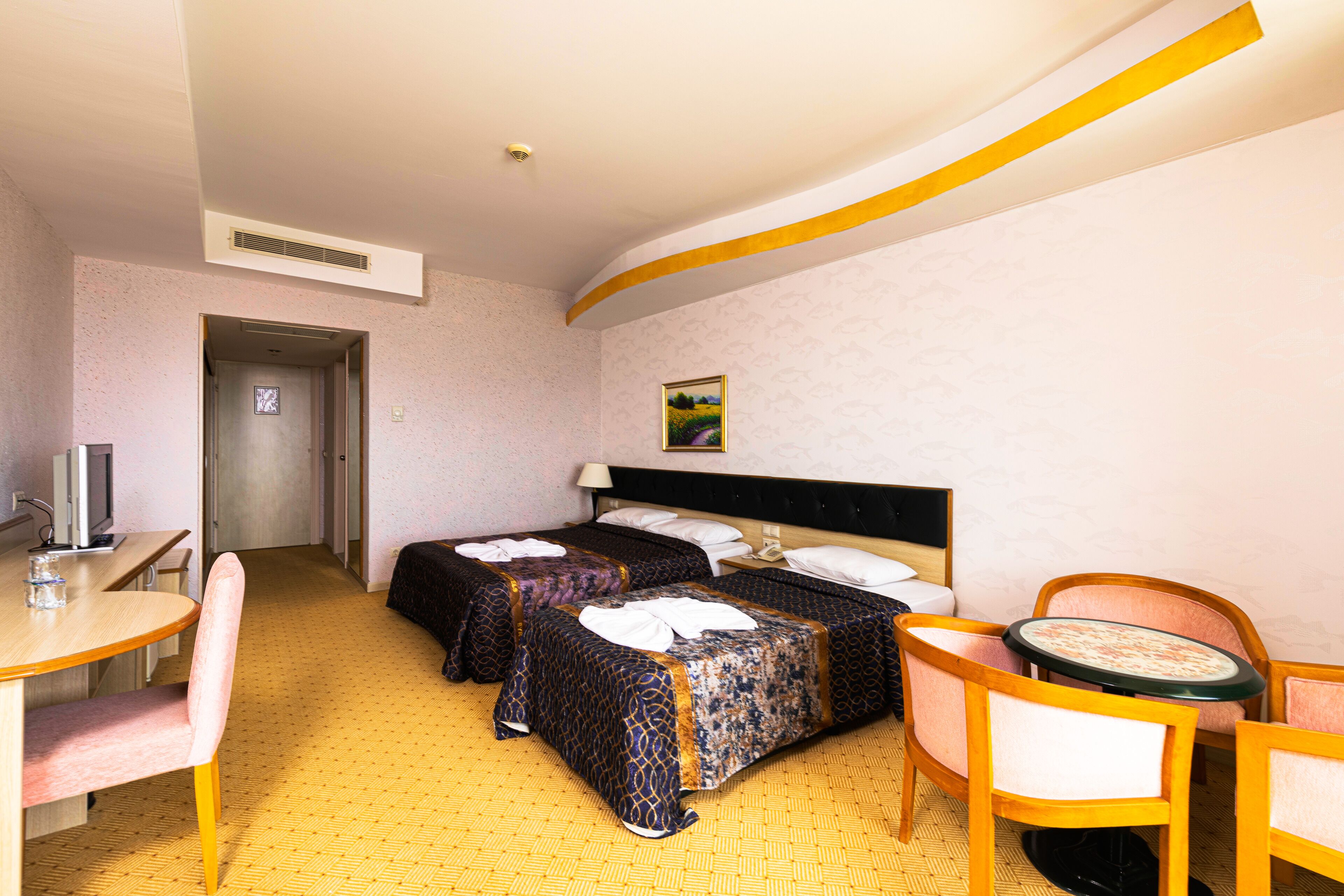A hotel room with two beds, a desk with a chair, a small table with a chair, and a painting on the wall.
