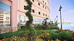 A hotel with a large glass facade, a well-maintained garden with orange flowers, and a view of a body of water. - Grand Cortez Halal Family Hotel - All Inclusive  (Alanya)