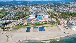 An aerial view of a coastal resort with a large sandy beach, clear blue waters, and a variety of buildings including a hotel with a pool and sun loungers. - Grand Cortez Halal Family Hotel - All Inclusive  (Alanya)