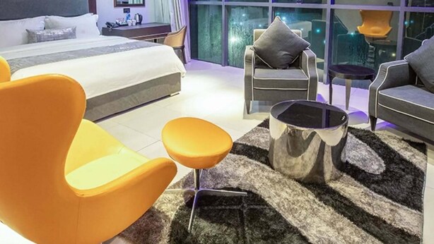 Een moderne hotelkamer met een groot bed, twee fauteuils, een bank, een salontafel en uitzicht op de stad door grote ramen.