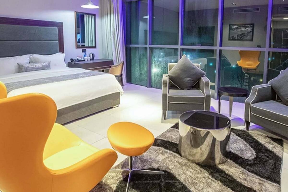 A modern hotel room with a large bed, two armchairs, a sofa, a coffee table, and a view of the city through large windows.