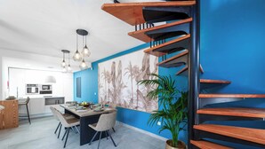 Una cocina moderna con armarios blancos, un comedor con mesa y sillas de madera, y una pared azul decorada con un gran mural tropical.