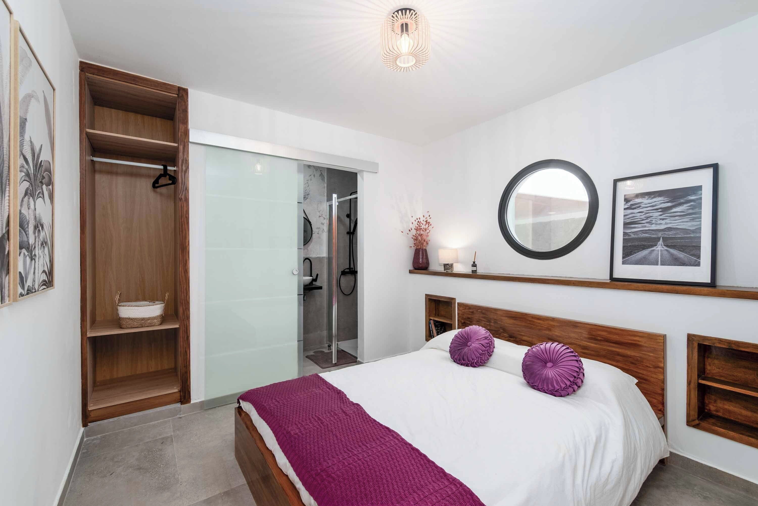 A bedroom with a bed, a wooden wardrobe, a glass shower enclosure, and a round mirror on the wall.